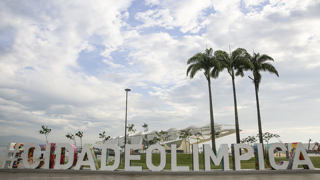 Na frente da imagem uma escultura interativa com a frase cidade olímpica com duas palmeiras atrás do lado direito e o Museu do Amanhã no plano de fundo