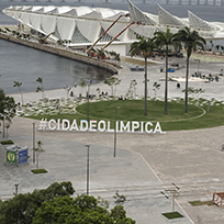 Na frente da imagem uma escultura interativa com a frase cidade olímpica com duas palmeiras atrás do lado direito e o Museu do Amanhã no plano de fundo