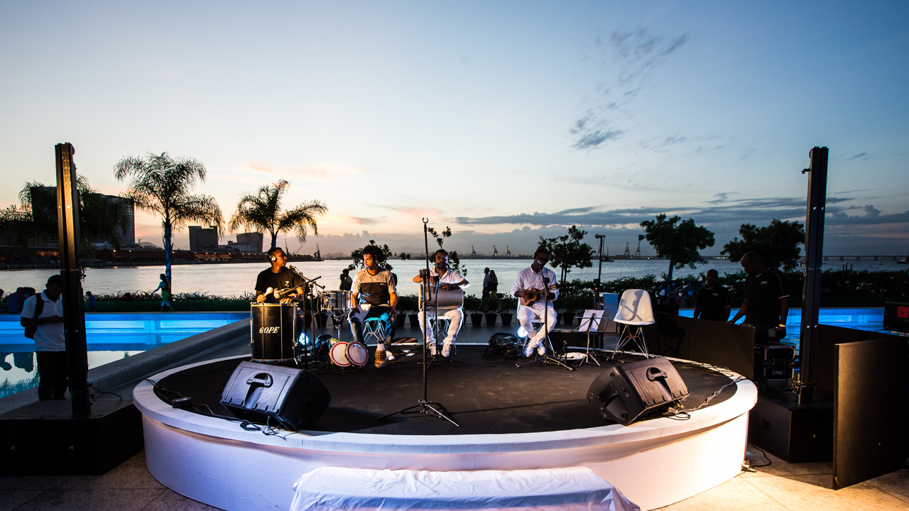 Foto feita na lateral do museu, com banda de música tocando em palco circular branco e, ao fundo, a Baía de Guanabara no pôr do sol.